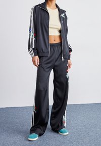 Svart satin träningsoverall med vita ränder, blommig broderi på ärmar och byxor, kort topp med rund halsringning, kombinerad med turkosa sneakers.