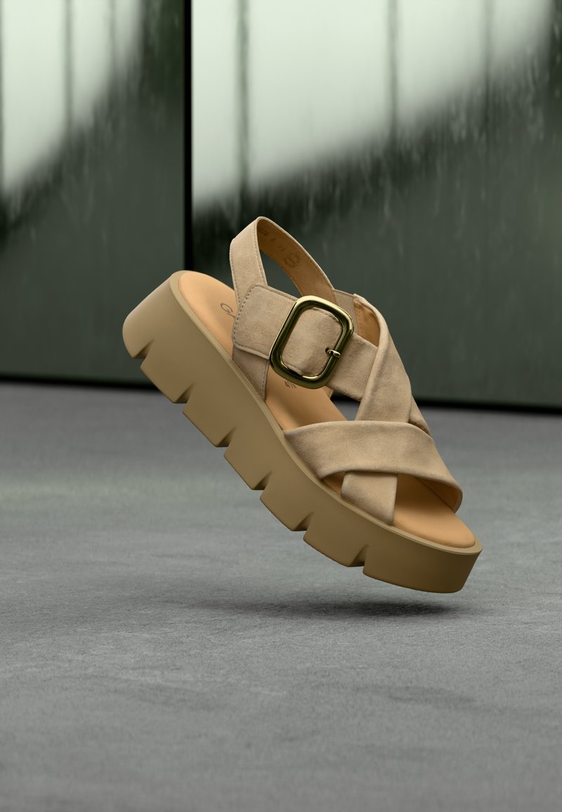 Beige cross-strap sandal with a chunky, ridged platform sole. Features a gold square buckle and soft suede-like texture.