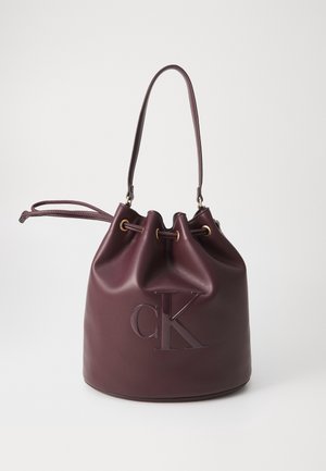 Sac seau en cuir violet foncé avec fermeture à cordon, bandoulière et logo CK embossé à l'avant sur fond uni.