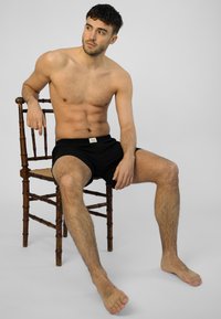 Černé boxerky na sedícím muži s krátkými vlasy. Má svalnatou postavu a je bosý, pózuje na dřevěné židli na jednobarevném pozadí.