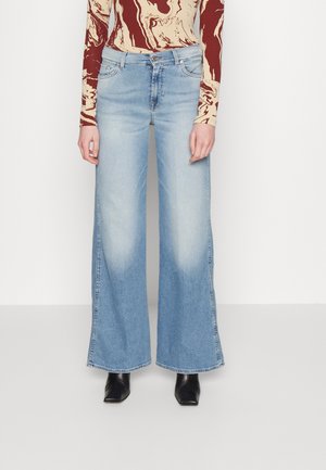 Osoba ubrana w jasnoniebieskie szerokie jeansy, czarne spiczaste buty i długorękawowy top z czerwonym i beżowym abstrakcyjnym wzorem, stojąca przy białej ścianie.