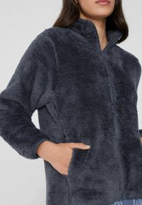 Chaqueta de forro polar gris oscuro difuso con cuello alto, cremallera frontal y bolsillos laterales. Textura suave y apariencia cálida, diseñada para la comodidad.