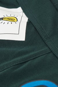 Tejido verde oscuro con textura visible presenta un parche gráfico blanco que representa una bombilla rodeada de líneas radiantes, cosido con acentos azules.