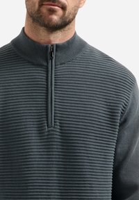 Man met een donkergrijze geribbelde gebreide trui met een korte rits en hoge kraag, waarbij de nek en onderzijde van het gezicht zichtbaar zijn.