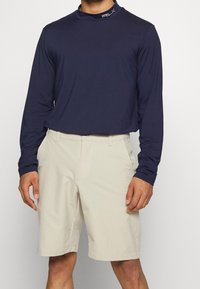 Man i marinblå långärmad skjorta instoppad i beige knälånga shorts med sidofickor, stående mot en enhetlig bakgrund.