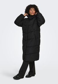 Mustassa toppatakissa on tikattu malli, korkea kaulus ja etutasku vetoketjulla. Takki on yllään mustien housujen ja kenkien päällä, ja siinä on väljä istuvuus sekä sivunapit.