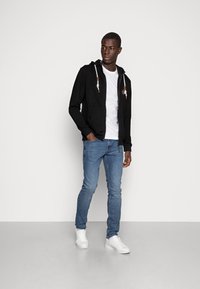 Sort hættejakke med lynlås og snoretræk samt brune detaljer, kombineret med en hvid T-shirt og blå jeans. Model iført hvide sneakers.