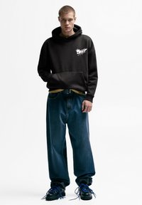 Črna kapucar z žepom spredaj in grafičnim detajlom, kombiniran z ohlapnimi modrimi jeans hlačami in črnimi škornji z modrimi vezalkami.