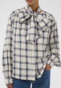 Blouse à carreaux beige, bleu et gris. Caractérisée par un grand col lavallière, une patte de boutonnage sur le devant et des épaules froncées. Manches longues avec poignets boutonnés.