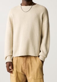 Maglione a coste beige con vestibilità rilassata, scollatura rotonda e maniche lunghe, abbinato a pantaloni marrone chiaro con tasche laterali.