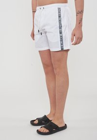 Costume da bagno bianco con dettagli del logo verticale nero, vita elastica e cordoncino. Abbinato a sandali neri da scivolo.