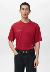 Tricou roșu din bumbac cu mâneci scurte, având text brodat alb. Asortat cu blugi negri din denim și o curea neagră.