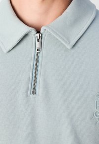 Ljusblå ribbad pikétröja med dragkedja i halsen, med en platt krage och broderad detalj på sidan. Mjukt material och åtsittande design.