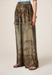 Weite Hosen aus einem mehrfarbigen Stoff mit einem Naturdruck aus Bäumen und Laub. Elastischer Bund und glatte Textur, ergänzt durch schwarze Sandalen.