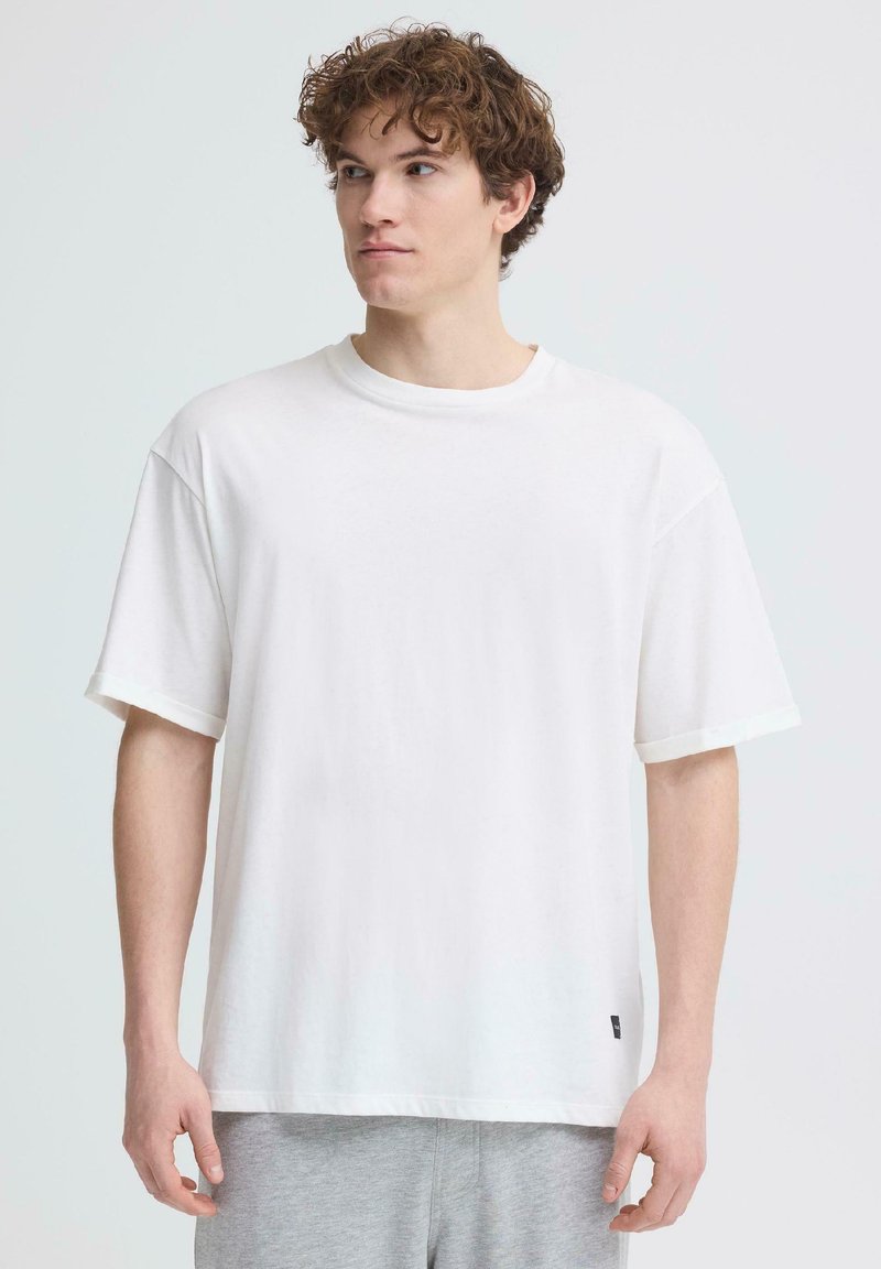 Jeune homme aux cheveux bruns bouclés, portant un t-shirt blanc uni oversize et un pantalon gris clair, regardant sur le côté.
