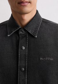 Close-up van een persoon die een zwart denim overhemd met zwarte knopen draagt en een geborduurd "Marc O'Polo" logo op de borst.