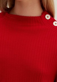Pull tricoté rouge à côtes avec un col montant, orné de deux boutons blancs sur l'épaule. Texture lisse, coupe ajustée.
