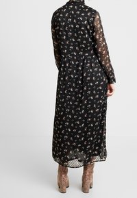 Svart blommig klänning med långa, genomskinliga ärmar; har ett prickigt mönster vid fållen. Tyget är lätt och har en avslappnad passform.