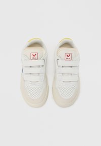 Valged tossud Velcro kinnitustega, perforatsioonidega varbale, heledate beežide aktsentidega, punase logoga keelel ja kollase detailiga kontsal.