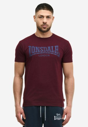 Mężczyzna w bordowej koszulce "Lonsdale London" i granatowych szortach z dopasowanym logo "Lonsdale London", stojący na białym tle.