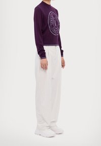 Pull en tricot violet avec un logo circulaire, associé à un pantalon large à rayures blanches et des baskets blanches. Tissu texturé et accents contrastés.