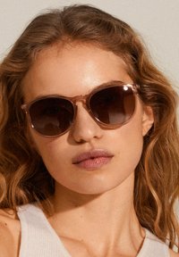 Sunglasses with a translucent pink frame and dark brown lenses. Rounded shape with subtle hardware accents at the temples.