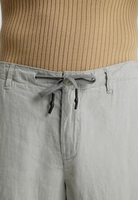 Jasnoszare lniane szorty mają elastyczny pasek ze sznurkiem zakończonym czarnymi plastikowymi końcówkami oraz kieszeniami po bokach, w luźnym kroju. Widoczny jest teksturowany prążkowany top powyżej.