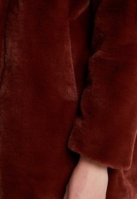 Bordowy płaszcz z futra syntetycznego o miękkiej teksturze, z luźnym krojem i lekko oversize'owymi rękawami. Gładka powierzchnia bez widocznych elementów metalowych.