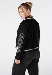 Blonde vrouw met een bun in haar haar draagt een zwarte jas met beige borduursels op de mouwen en onderkant, gecombineerd met donkergrijze spijkerbroek, zijwaarts gekeerd.