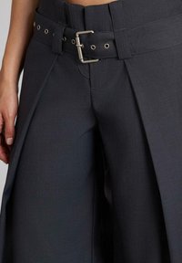 Pantalones de tiro alto en gris oscuro con un cinturón ancho, que incluye una hebilla plateada y ojales de metal, y un diseño geométrico entallado.