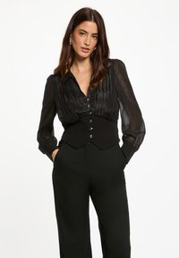 Blusa nera con colletto e maniche lunghe plissettate e trasparenti, con chiusura a bottoni sul davanti. Tessuto testurizzato con dettagli lucidi, abbinata a pantaloni neri su misura.