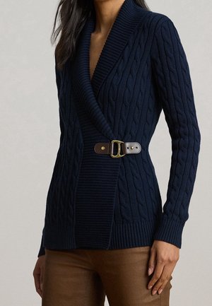 Femme portant un cardigan en maille torsadée bleu marine avec un col châle et une ceinture en cuir marron avec une boucle dorée à la taille.