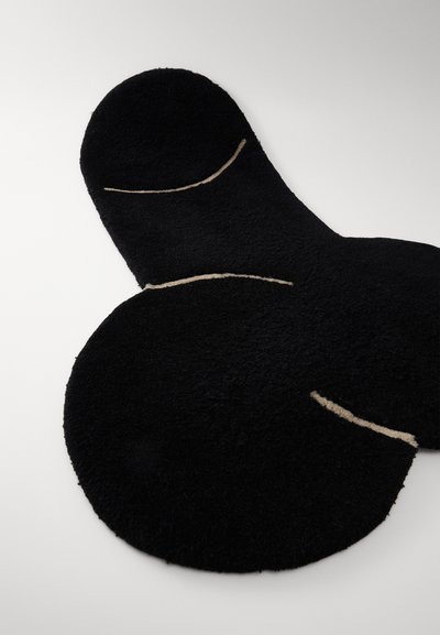 Zwarte, plush vloerkleed in de vorm van een gestileerde anatomische figuur, met crèmekleurige lijnen die het ontwerp accentueren. Zachte textuur, onregelmatige vorm.