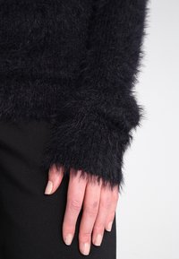 Czarny puszysty sweter z długimi rękawami, o miękkiej fakturze i lekko oversize'owych mankietach. Zbliżenie na rękę opierającą się na czarnej powierzchni.
