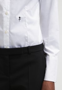 Camisa blanca de botones con tejido texturizado, pequeño bordado de rosa negra en el lado izquierdo, combinada con pantalones negros a medida.