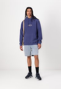 Fialová mikina s logom, skombinovaná so svetlými džínsovými šortkami. Čierne ponožky a športové topánky dopĺňajú outfit. Model stojí na bielom pozadí.