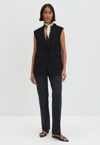 Schwarzer ärmelloser Blazer mit scharfen Revers und Seitentaschen, kombiniert mit dunklen Hosen und einem leichten Schal, der am Hals gebunden ist. Flache Sandalen runden den Look ab.