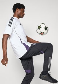 Witte korte mouw shirt met zwarte strepen, zwarte broek met paarse accenten en zwarte sportschoenen. Een voetbal zweeft nabij de knie.