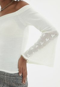 Weißes Off-Shoulder-Oberteil aus strukturiertem Strickstoff mit ausgestellten, durchsichtigen Ärmeln, die herzförmige Ausschnitte aufweisen. Mit einer dekorativen Kettenakzent versehen.