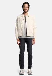 Man met krullend haar in een crèmekleurige knopenjacket, grijze shirt, zwarte jeans en witte sneakers, staande tegen een effen witte achtergrond.