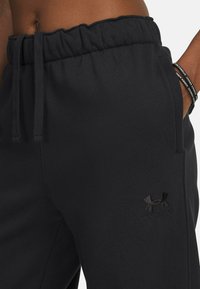 Pantalons de jogging noirs avec une taille élastique et un cordon de serrage, arborant un logo Under Armour noir brodé sur la cuisse gauche. Texture douce.