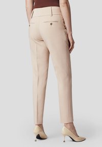 Beżowe spodnie o dopasowanym kroju z gładką fakturą, wąskie, z dekoracyjnymi guzikami z tyłu. Noszone z beżowymi butami na obcasie.