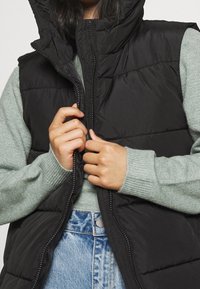 Svart vadderad väst med hög krage, dragkedja fram och quiltad textur, bärs över en ljusgrön tröja och blåa denimjeans.