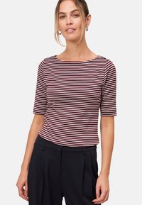 Vrouw met lichtbruin haar, gekleed in zilveren oorbellen, een maroon en witte gestreepte bootneck top en zwarte hoge taillebroeken tegen een witte achtergrond.