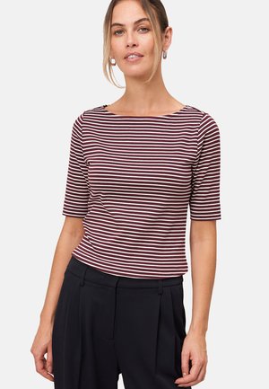 Femme aux cheveux châtain clair portant des boucles d'oreilles en argent, un haut à col bateau maroon et blanc rayé, et un pantalon noir taille haute, devant un fond blanc.