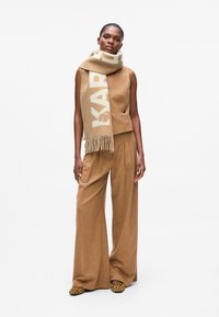 Brauner Wollweste und weit geschnittene Hose, kombiniert mit einem gestreiften Schal in Beige und Creme, der Text enthält; Tierdruckschuhe runden den Look ab.