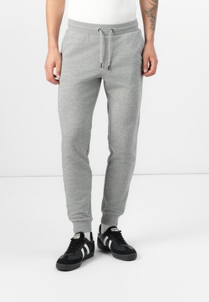 Mand iført lysegrå joggingbukser med snøre og sorte sneakers med hvide striber, stående mod en ensfarvet hvid baggrund.