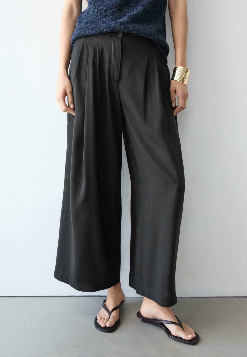 Personne portant un pantalon large noir, un haut sans manches bleu marine, un bracelet en or et des sandales noires à enfiler, debout contre un mur blanc.