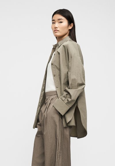 Νεαρή γυναίκα με υπερμεγέθη μπεζ πουκάμισο πάνω από λευκό τοπ και φαρδιά παντελόνια ταυπέ, στέκεται πλευρικά μπροστά σε απλό φόντο.