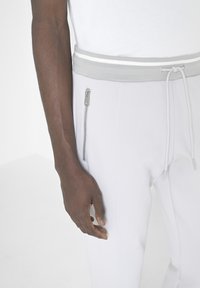 Joggers gris claro con una cintura acanalada que presenta una franja blanca, complementados con un bolsillo lateral con cremallera, mostrados en una mano.